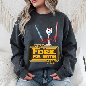 May include: A dark gray crewneck sweatshirt featuring a graphic of the character Forky holding a blue and a red lightsaber. Below the character, the yellow text reads: MAY THE FORK BE WITH YOU, styled in a classic science fiction movie font.