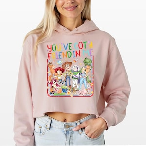 May include: A light pink cropped hoodie featuring a colorful graphic of various animated characters from the Toy Story franchise. The text "YOU'VE GOT A FRIEND IN ME" is printed in multi-colored letters above the group of characters.