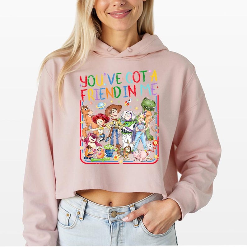 May include: A light pink cropped hoodie featuring a colorful graphic of various animated characters from the Toy Story franchise. The text "YOU'VE GOT A FRIEND IN ME" is printed in multi-colored letters above the group of characters.