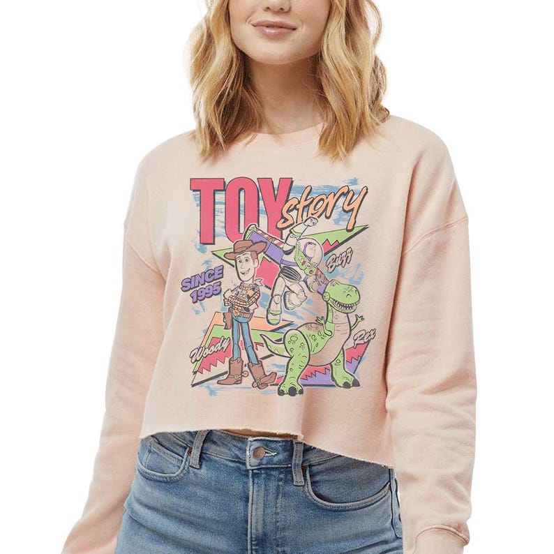 May include: A light peach-colored long-sleeve crop sweatshirt featuring a graphic print of Toy Story characters Woody, Buzz Lightyear, and Rex. The design includes the text "TOY story", "SINCE 1995", "Woody", "Buzz", and "Rex" in a colorful retro style.