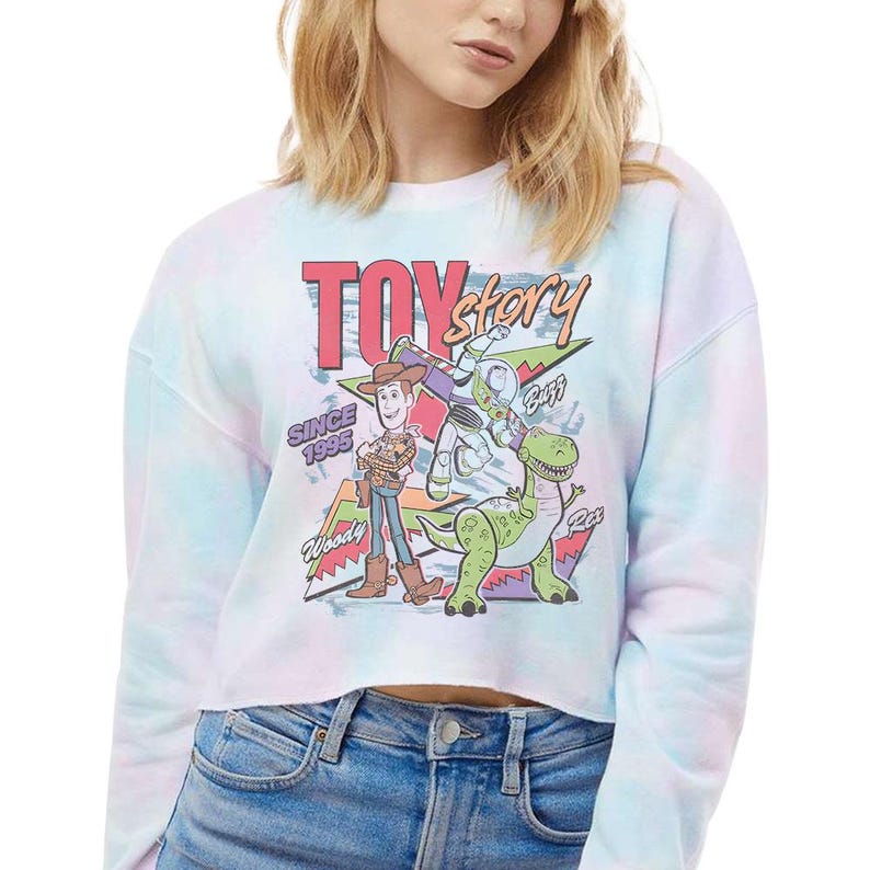 May include: A long-sleeve cropped sweatshirt in a pastel tie-dye pattern featuring a Toy Story graphic. The print displays Woody, Buzz Lightyear, and Rex with the text Toy Story, Since 1995, Woody, Buzz, and Rex in a colorful, retro-inspired design.