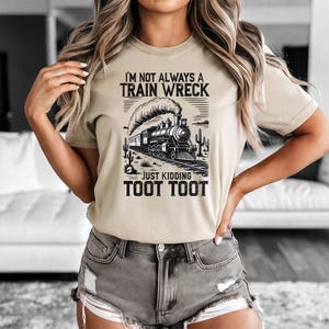 May include: Beige t-shirt with a black and white graphic of a vintage train, desert landscape, and the text "I'M NOT ALWAYS A TRAIN WRECK JUST KIDDING TOOT TOOT". The shirt has short sleeves and a crew neck.