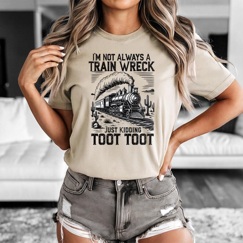 May include: Beige t-shirt with a black and white graphic of a vintage train, desert landscape, and the text "I'M NOT ALWAYS A TRAIN WRECK JUST KIDDING TOOT TOOT". The shirt has short sleeves and a crew neck.