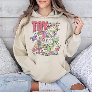May include: A beige hooded sweatshirt featuring a graphic print of the Toy Story characters Woody, Buzz Lightyear, and Rex. The design includes the text "TOY story" at the top, "SINCE 1995" on the left, and the names "Woody", "Buzz", and "Rex" near each character.