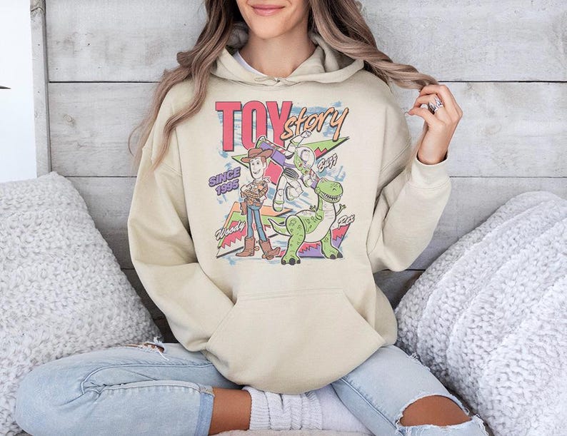May include: A beige hooded sweatshirt featuring a graphic print of the Toy Story characters Woody, Buzz Lightyear, and Rex. The design includes the text "TOY story" at the top, "SINCE 1995" on the left, and the names "Woody", "Buzz", and "Rex" near each character.