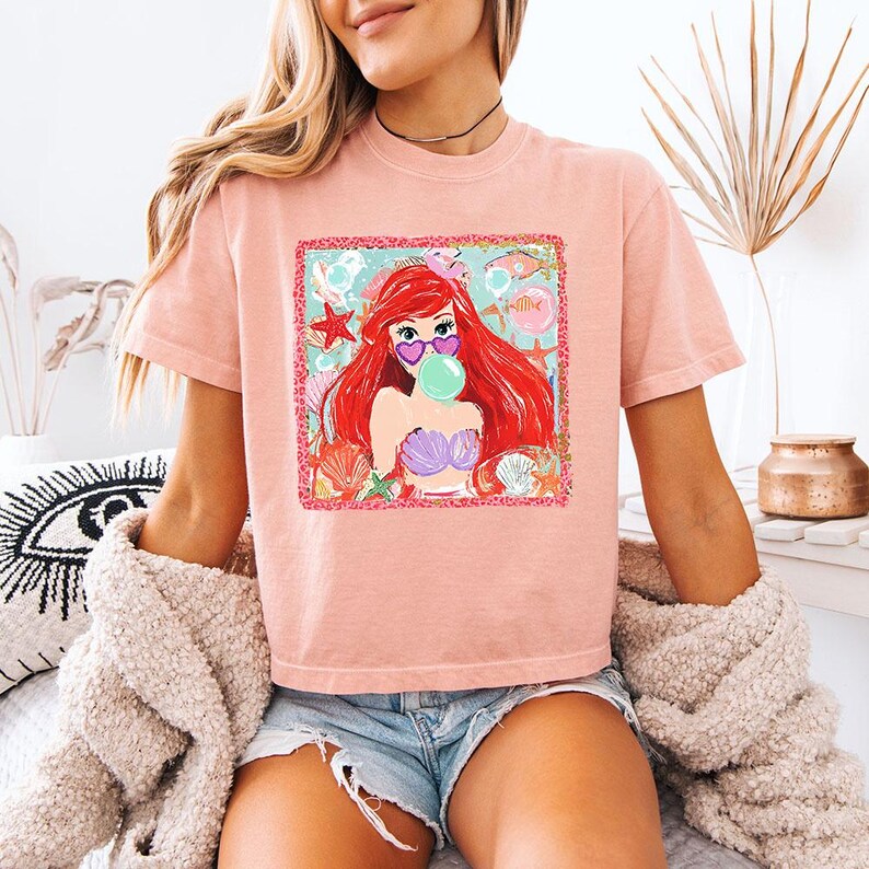 K&ouml;nnte beinhalten: Hellrosa T-Shirt mit einem farbenfrohen Aufdruck einer Cartoon-Meerjungfrau mit roten Haaren, die eine herzf&ouml;rmige Sonnenbrille tr&auml;gt und eine Blase bl&auml;st. Die Grafik enth&auml;lt Muscheln, Seesterne und Fische. Das T-Shirt wird mit Jeansshorts getragen.