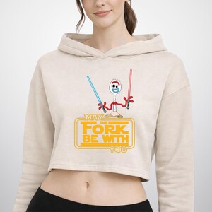 Puede incluir: Una sudadera corta de color beige con el gr&aacute;fico de un personaje tipo spork blanco con brazos de limpiapipas rojos que sostiene un sable de luz azul y otro rojo. Debajo, el texto amarillo dice MAY THE FORK BE WITH YOU en fuente bloque.