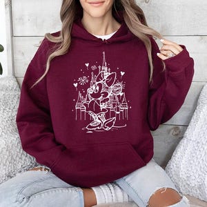 May include: Burgundy hoodie with a white graphic of a cartoon character in front of a castle. The hoodie has a drawstring hood and a front pocket. The graphic includes fireworks and hearts. The person is wearing light blue jeans.