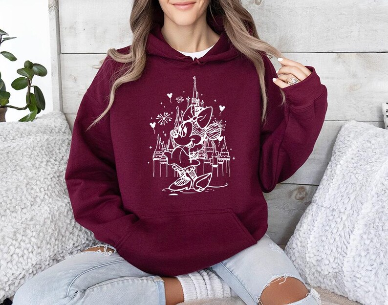 May include: Burgundy hoodie with a white graphic of a cartoon character in front of a castle. The hoodie has a drawstring hood and a front pocket. The graphic includes fireworks and hearts. The person is wearing light blue jeans.