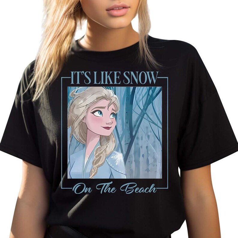 May include: Black t-shirt with a white frame around a cartoon image of Elsa from Frozen. The text "It's Like Snow - On The Beach" is printed on the shirt.