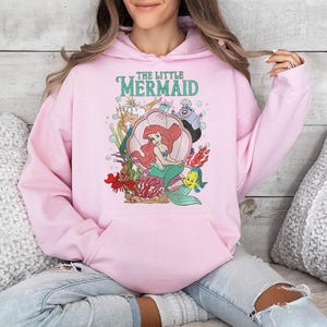 May include: A light pink hooded sweatshirt featuring a graphic print of The Little Mermaid characters including Ariel, King Triton, Ursula, and Flounder. The design includes the text The Little Mermaid in teal lettering above the characters.