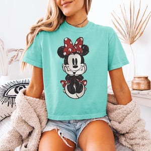 Puede incluir: Una camiseta de color turquesa de manga corta con un gr&aacute;fico central de Minnie Mouse con un lazo rojo de lunares blancos, orejas negras y un estampado de estilo vintage desgastado. La camiseta tiene cuello redondo y corte holgado.