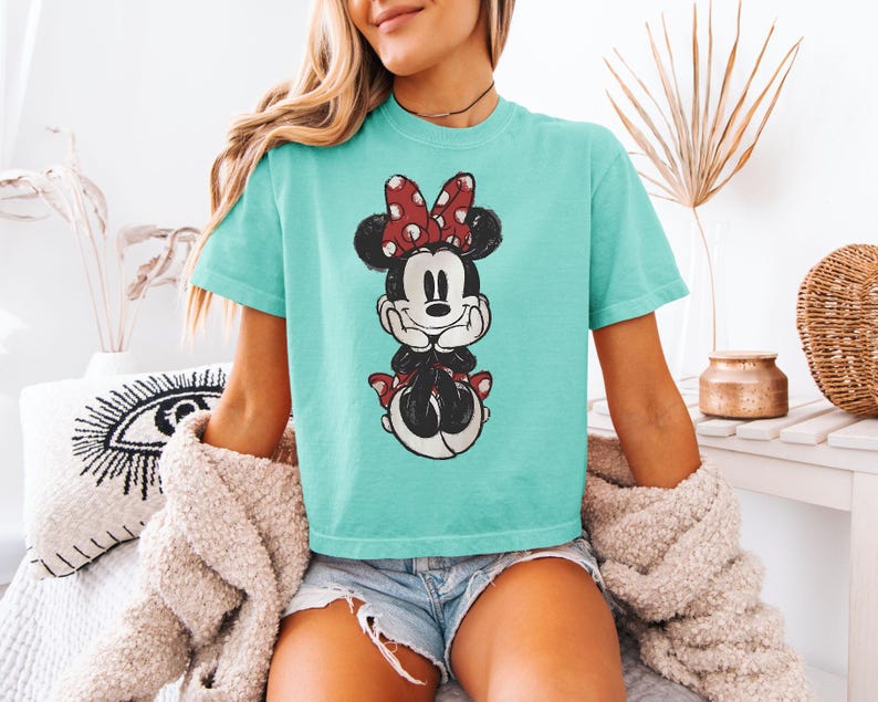 Puede incluir: Una camiseta de color turquesa de manga corta con un gr&aacute;fico central de Minnie Mouse con un lazo rojo de lunares blancos, orejas negras y un estampado de estilo vintage desgastado. La camiseta tiene cuello redondo y corte holgado.