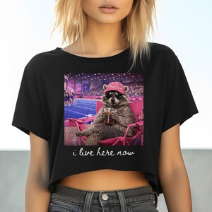 May include: Black crop top featuring a graphic of a raccoon wearing sunglasses and a pink hat, seated in a pink chair, holding a drink. The image has the text "i live here now" in white.