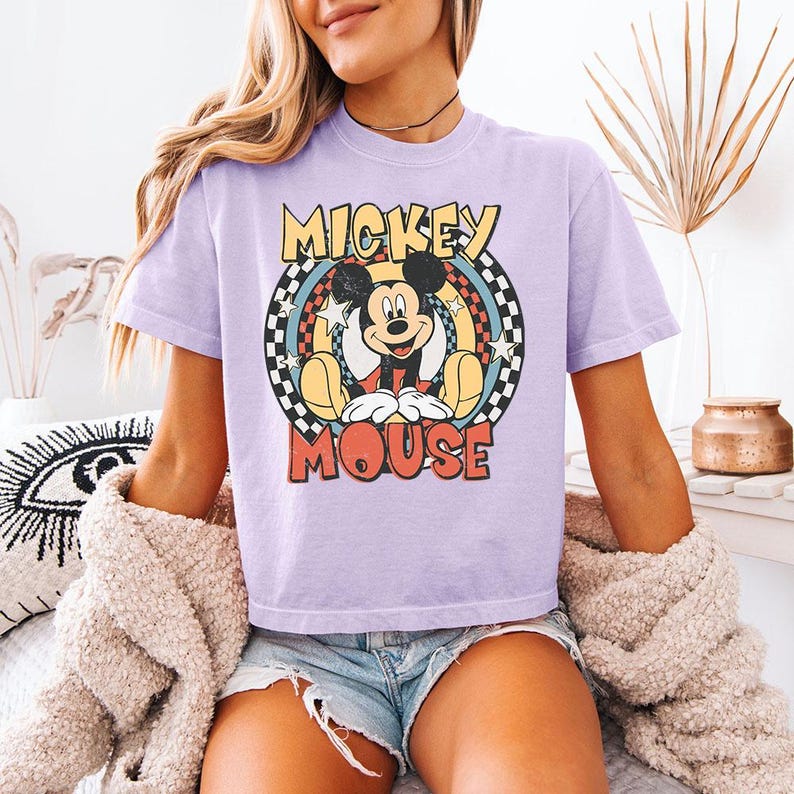 Pu&ograve; includere: Una t-shirt a maniche corte color viola chiaro con una grafica di Mickey Mouse centrata all'interno di un design circolare. La grafica include il testo MICKEY MOUSE in grassetto, circondato da un bordo a scacchi, anelli colorati e stelle bianche.