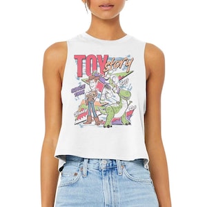 Puede incluir: Un top corto sin mangas de color blanco con un estampado gr&aacute;fico colorido de los personajes Woody, Buzz Lightyear y Rex de Toy Story. El dise&ntilde;o incluye el texto "TOY story", "SINCE 1995", "Woody", "Buzz" y "Rex" en varias fuentes y colores.