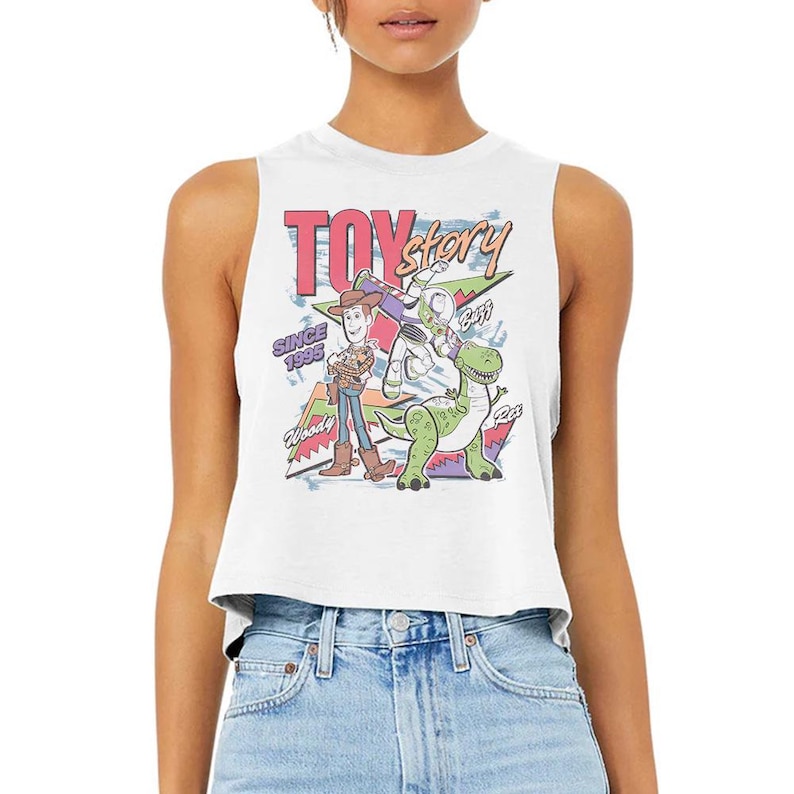 Puede incluir: Un top corto sin mangas de color blanco con un estampado gr&aacute;fico colorido de los personajes Woody, Buzz Lightyear y Rex de Toy Story. El dise&ntilde;o incluye el texto "TOY story", "SINCE 1995", "Woody", "Buzz" y "Rex" en varias fuentes y colores.