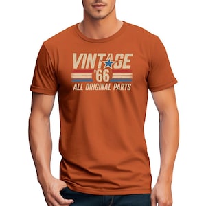 Puede incluir: Una camiseta de color &oacute;xido con el texto "VINTAGE '66 ALL ORIGINAL PARTS" en un estilo retro desgastado. El dise&ntilde;o incluye una estrella azul y rayas rojas y azules. La camiseta es de manga corta.