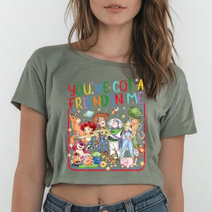 May include: An olive green cropped t-shirt featuring a colorful graphic print of various animated toy characters. The text "YOU'VE GOT A FRIEND IN ME" is printed in a rainbow-colored font above the characters, all enclosed within a red rectangular border.