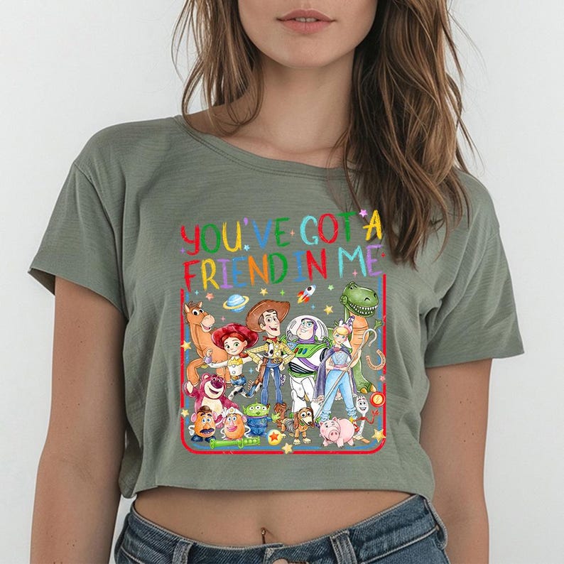 May include: An olive green cropped t-shirt featuring a colorful graphic print of various animated toy characters. The text "YOU'VE GOT A FRIEND IN ME" is printed in a rainbow-colored font above the characters, all enclosed within a red rectangular border.