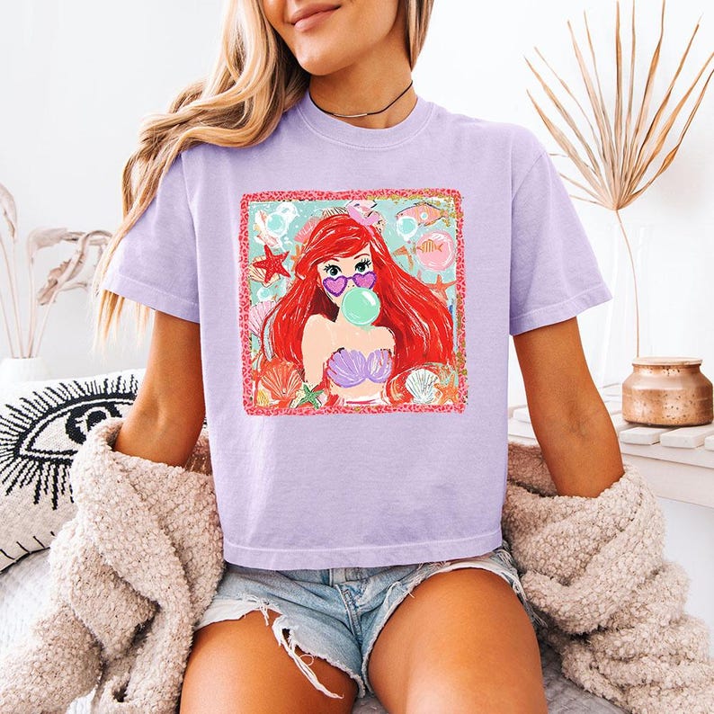 K&ouml;nnte beinhalten: Helllila T-Shirt mit einem Aufdruck einer Meerjungfrau mit roten Haaren, die eine herzf&ouml;rmige Sonnenbrille tr&auml;gt und eine Blase bl&auml;st. Der Aufdruck ist von Muscheln und Seesternen umgeben. Das T-Shirt hat kurze &Auml;rmel.