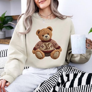 May include: Beige sweatshirt featuring a brown teddy bear design wearing a sweater with a logo. The teddy bear has a detailed, textured appearance. The sweatshirt is a neutral color, suitable for casual wear.