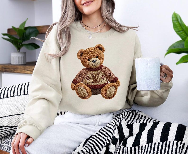 May include: Beige sweatshirt featuring a brown teddy bear design wearing a sweater with a logo. The teddy bear has a detailed, textured appearance. The sweatshirt is a neutral color, suitable for casual wear.