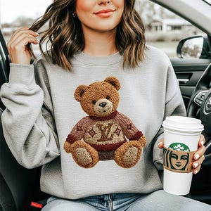 May include: A light gray sweatshirt featuring a brown teddy bear wearing a sweater with a logo design. The teddy bear has a smiling face and is sitting on the front of the sweatshirt. A white coffee cup with a brown sleeve is held in the hand.