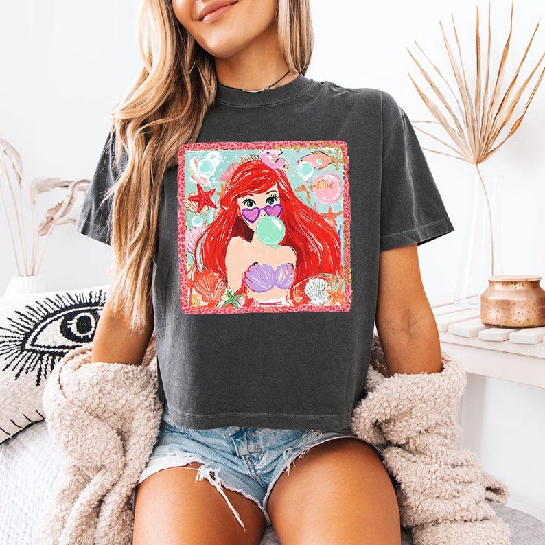 K&ouml;nnte beinhalten: Dunkelgraues T-Shirt mit einem farbenfrohen Aufdruck einer Cartoon-Meerjungfrau mit roten Haaren, herzf&ouml;rmiger Sonnenbrille und einer Blase. Die Grafik ist von Muscheln und Seesternen umgeben. Das Shirt ist ein l&auml;ssiger, bequemer Stil.