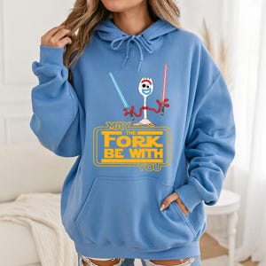 Op de afbeelding: Een lichtblauwe hoodie met een afbeelding van het personage Forky die een blauw en een rood lichtzwaard vasthoudt. Onder het personage staat de gele tekst: MAY THE FORK BE WITH YOU. Het kledingstuk heeft een kangoeroezak en trekkoorden.