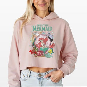 Puede incluir: Una sudadera corta de color rosa claro con un estampado gr&aacute;fico de los personajes de La Sirenita, incluyendo a Ariel, el Rey Trit&oacute;n, &Uacute;rsula, Sebasti&aacute;n y Flounder. El dise&ntilde;o incluye burbujas y elementos submarinos con el texto The Little Mermaid.