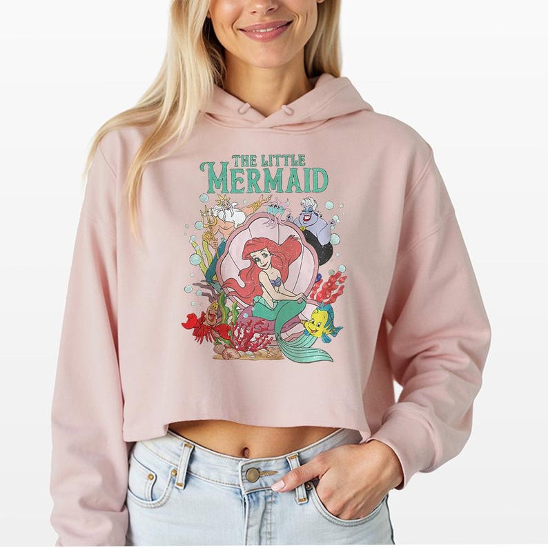 Puede incluir: Una sudadera corta de color rosa claro con un estampado gr&aacute;fico de los personajes de La Sirenita, incluyendo a Ariel, el Rey Trit&oacute;n, &Uacute;rsula, Sebasti&aacute;n y Flounder. El dise&ntilde;o incluye burbujas y elementos submarinos con el texto The Little Mermaid.