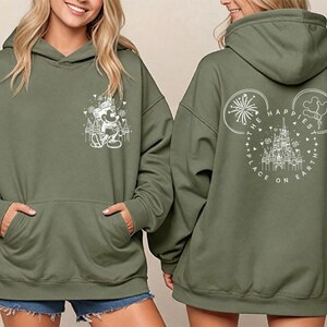 Pu&ograve; includere: Felpa con cappuccio verde oliva con tasca frontale e cappuccio. La parte anteriore presenta una grafica bianca di Mickey Mouse. La parte posteriore ha una grafica bianca di orecchie di Mickey Mouse con fuochi d'artificio e il testo "The Happiest Place on Earth".