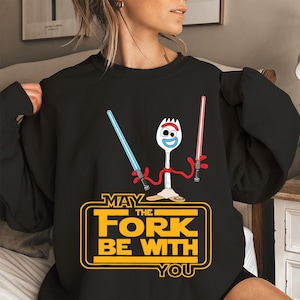 May include: A black long-sleeve sweatshirt featuring a graphic of a white plastic spork character with red pipe cleaner arms holding a blue and a red lightsaber. Below the character, yellow text reads MAY THE FORK BE WITH YOU.