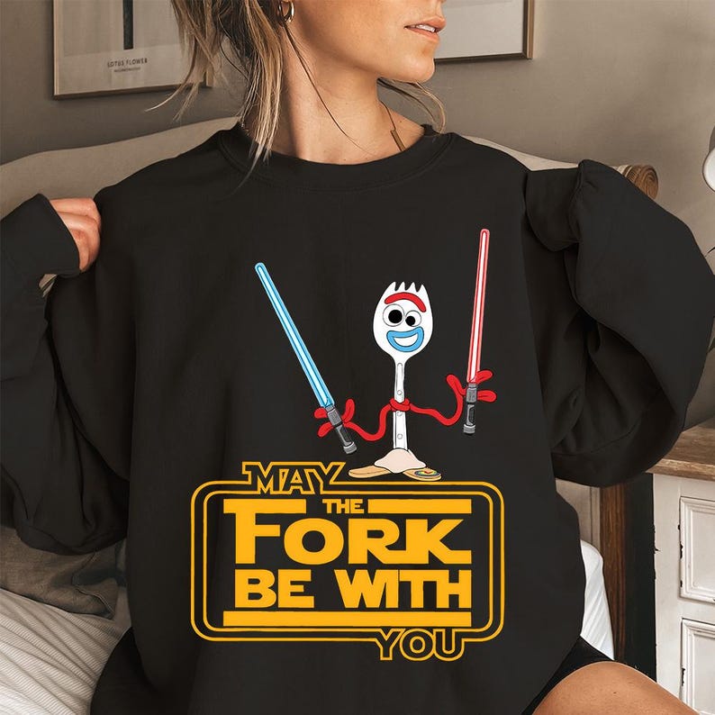 May include: A black long-sleeve sweatshirt featuring a graphic of a white plastic spork character with red pipe cleaner arms holding a blue and a red lightsaber. Below the character, yellow text reads MAY THE FORK BE WITH YOU.