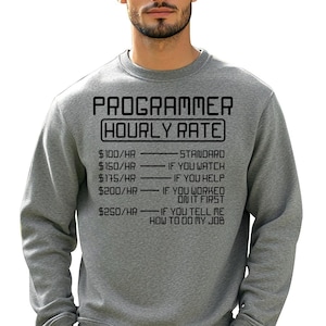 Può includere: Una felpa grigia con la scritta nera "PROGRAMMER HOURLY RATE". Il testo elenca tariffe orarie da $100 a $250 con condizioni umoristiche come "Se guardi" e "Se mi dici come fare il mio lavoro."