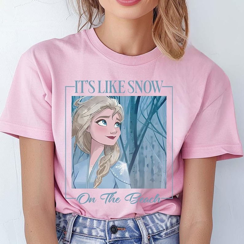 May include: Pink t-shirt with a graphic of Elsa from Frozen. The graphic features Elsa in a blue dress with a white trim and blonde hair. The text on the shirt reads "It's Like Snow On The Beach".