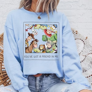Puede incluir: Sudadera azul claro con un gr&aacute;fico enmarcado de personajes de Toy Story. La imagen incluye a Woody, Buzz Lightyear y otros personajes. El texto "YOU'VE GOT A FRIEND IN ME" est&aacute; impreso debajo.