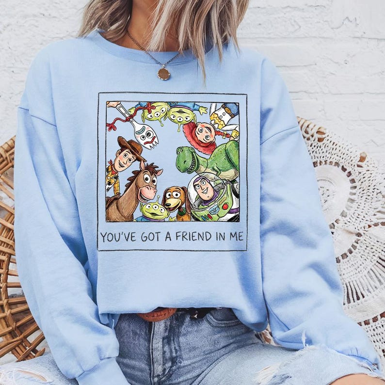 Puede incluir: Sudadera azul claro con un gr&aacute;fico enmarcado de personajes de Toy Story. La imagen incluye a Woody, Buzz Lightyear y otros personajes. El texto "YOU'VE GOT A FRIEND IN ME" est&aacute; impreso debajo.