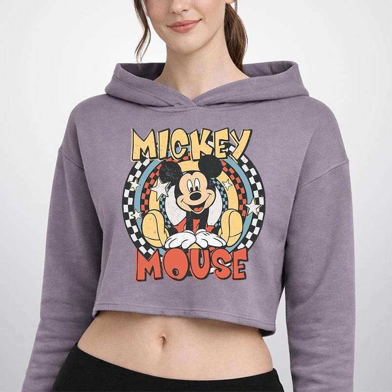 May include: A mauve long-sleeve cropped hoodie featuring a graphic print of Mickey Mouse. The design includes the text MICKEY MOUSE in yellow and orange, surrounded by a circular checkered border with stars and rainbow-colored concentric rings.