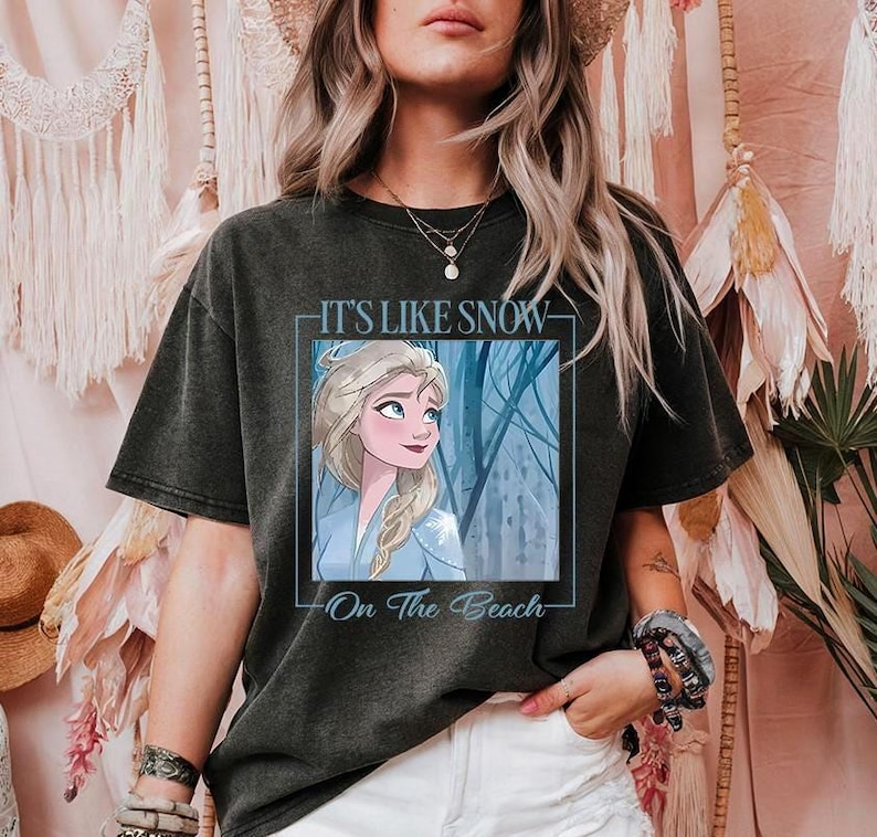 May include: A black t-shirt with a graphic of a cartoon character with blonde hair and blue eyes. The text on the shirt reads "It's Like Snow On The Beach".