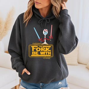 Op de afbeelding: Een donkergrijze hoodie met een afbeelding van een wit spork-personage dat een blauw en een rood lichtzwaard vasthoudt. Daaronder staat de gele tekst MAY THE FORK BE WITH YOU, geplaatst in een rechthoekig kader met afgeronde hoeken.