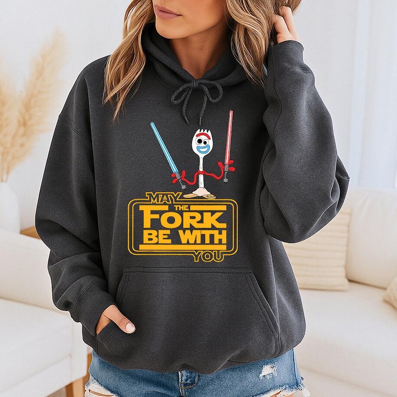 Op de afbeelding: Een donkergrijze hoodie met een afbeelding van een wit spork-personage dat een blauw en een rood lichtzwaard vasthoudt. Daaronder staat de gele tekst MAY THE FORK BE WITH YOU, geplaatst in een rechthoekig kader met afgeronde hoeken.
