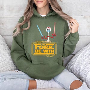 Op de afbeelding: Een olijfgroene hoodie met een afbeelding van een witte spork die een blauw en een rood lichtzwaard vasthoudt. Onder het personage staat de gele tekst "MAY THE FORK BE WITH YOU" binnen een rechthoekig kaderontwerp.