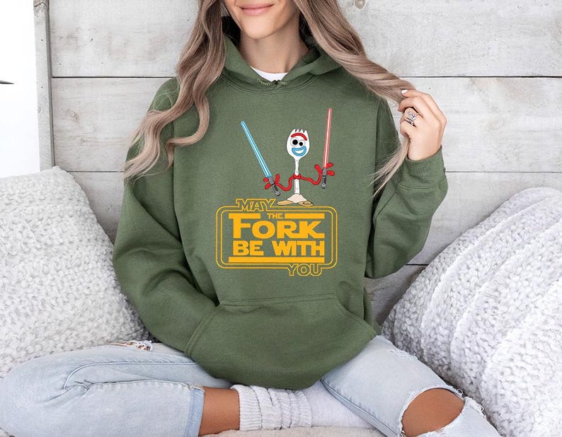 Op de afbeelding: Een olijfgroene hoodie met een afbeelding van een witte spork die een blauw en een rood lichtzwaard vasthoudt. Onder het personage staat de gele tekst "MAY THE FORK BE WITH YOU" binnen een rechthoekig kaderontwerp.