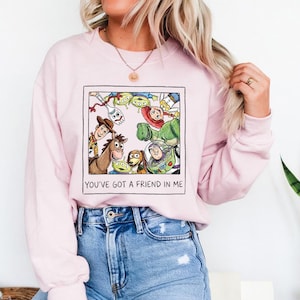 Puede incluir: Una sudadera rosa claro con un gr&aacute;fico enmarcado de personajes de Toy Story. El gr&aacute;fico incluye a Woody, Buzz Lightyear y otros personajes. El texto "YOU'VE GOT A FRIEND IN ME" est&aacute; impreso debajo.