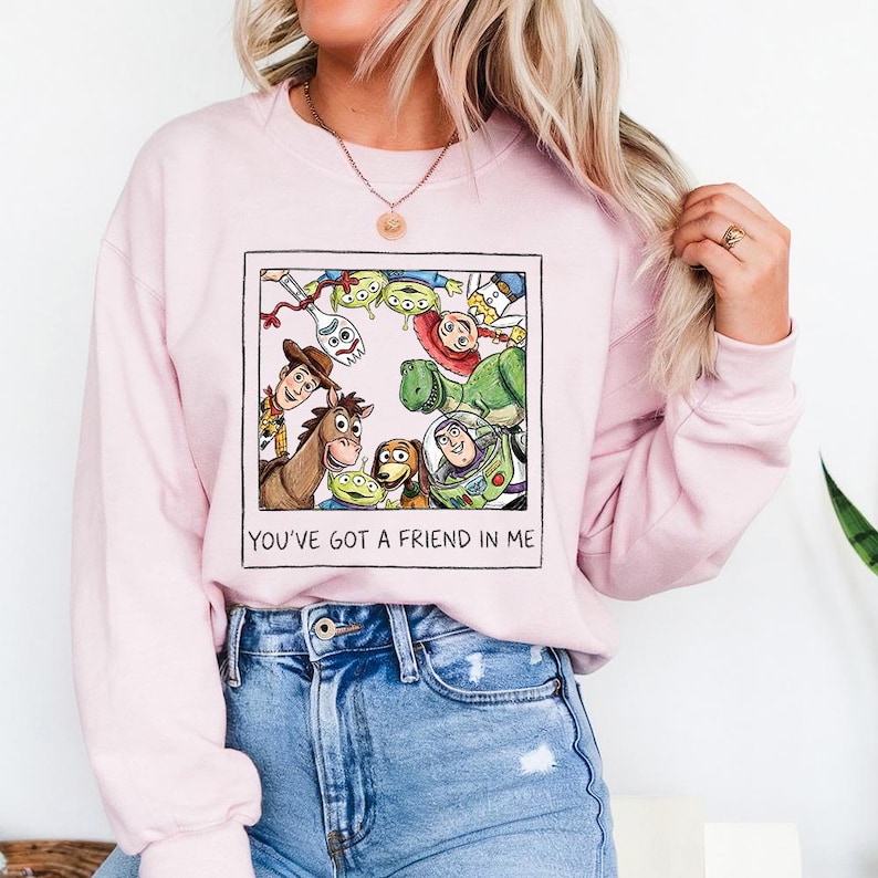 Puede incluir: Una sudadera rosa claro con un gr&aacute;fico enmarcado de personajes de Toy Story. El gr&aacute;fico incluye a Woody, Buzz Lightyear y otros personajes. El texto "YOU'VE GOT A FRIEND IN ME" est&aacute; impreso debajo.
