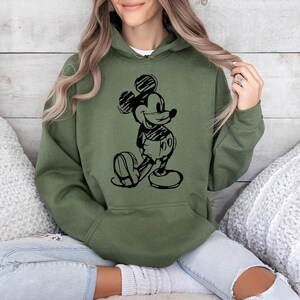 May include: An olive green hoodie featuring a black sketch of Mickey Mouse. The hooded sweatshirt has a front pocket and a drawstring hood. The graphic shows Mickey Mouse in a classic pose, with his hands behind his back.