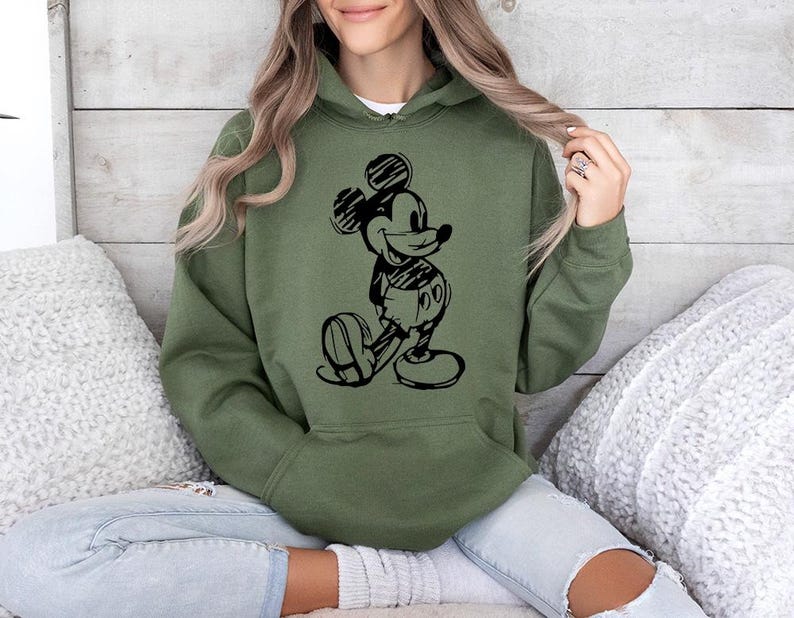 May include: An olive green hoodie featuring a black sketch of Mickey Mouse. The hooded sweatshirt has a front pocket and a drawstring hood. The graphic shows Mickey Mouse in a classic pose, with his hands behind his back.