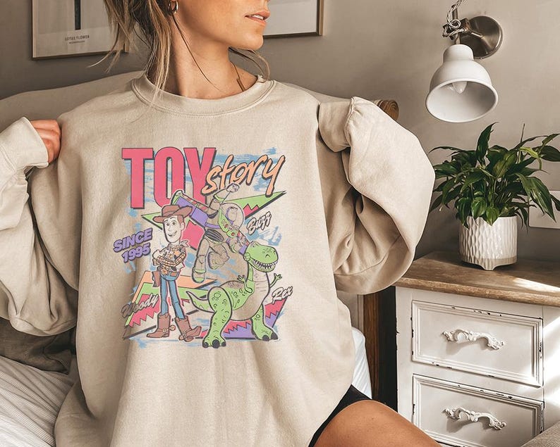 May include: A beige crewneck sweatshirt featuring a colorful graphic print of Toy Story characters Woody, Buzz Lightyear, and Rex. The design includes the text Toy Story, Since 1995, Woody, Buzz, and Rex, set against a background of geometric shapes.